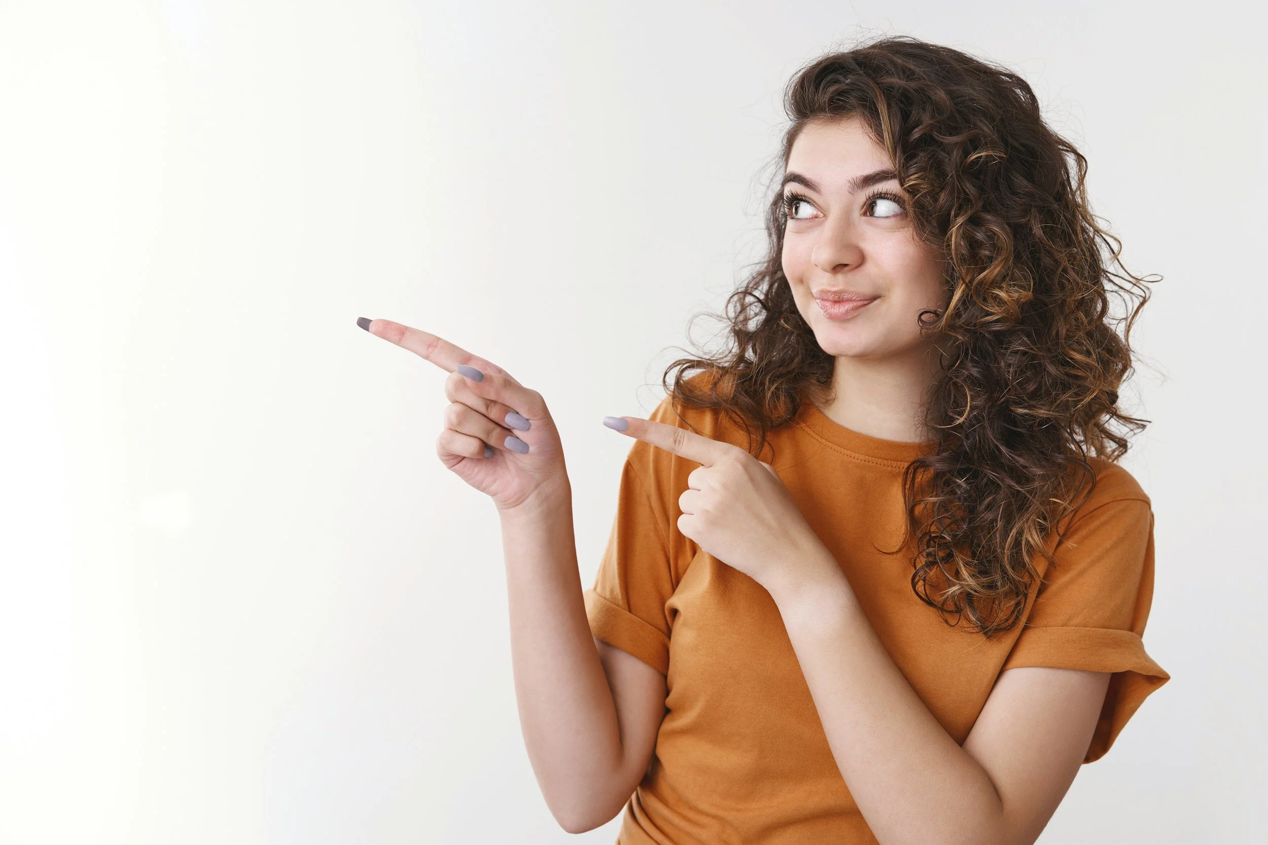 Young woman with curly brown hair and light skin pointing to the left, smiling, wearing a brown shirt, standing against a plain white background.
