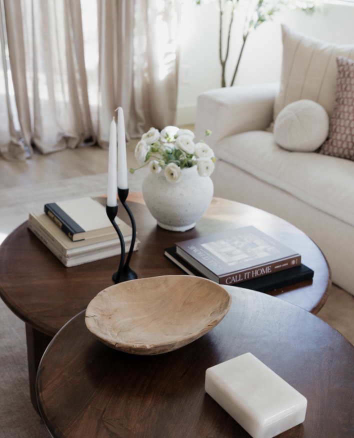 Living room with two wooden coffee tables, a white vase with white flowers, candles, books, and a white couch with cushions.