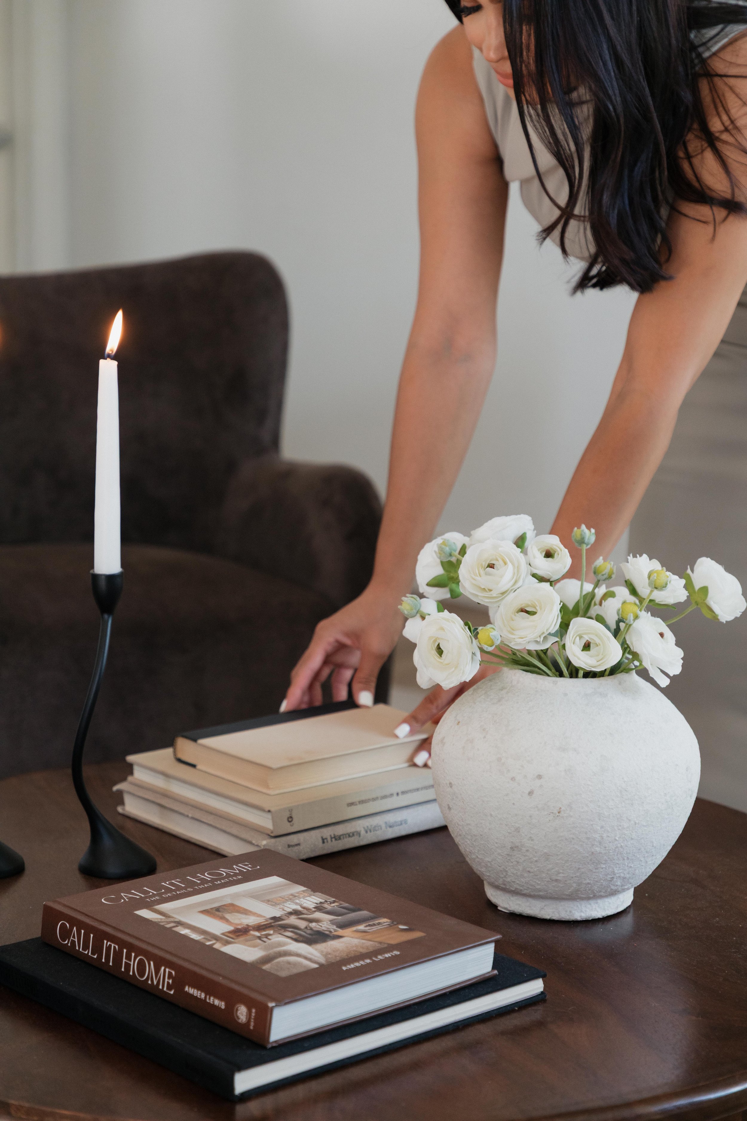 A woman is arranging flowers on a table with books, a white vase, and a lit candle nearby.
