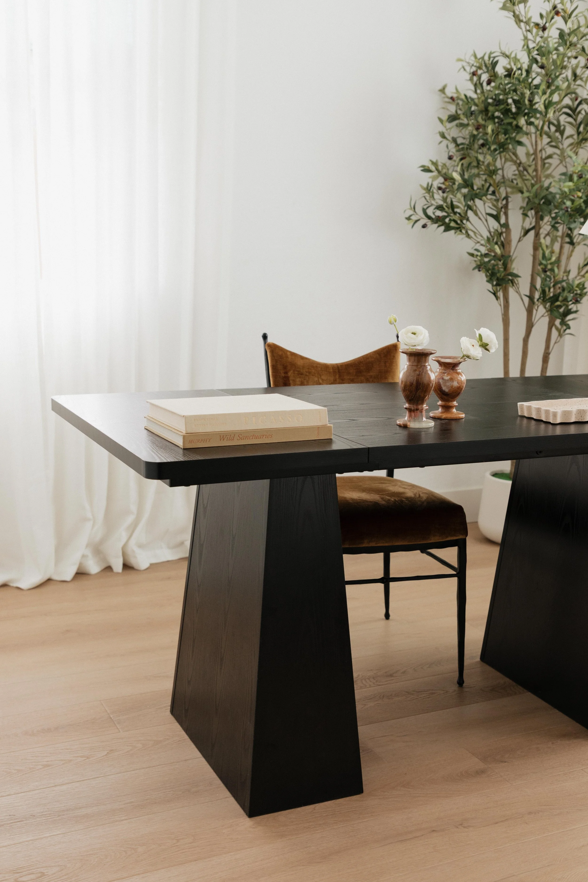 A sophisticated office with a black wooden desk, a brown chair, two small vases with white flowers, a stack of two books, and a rectangular decorative item.