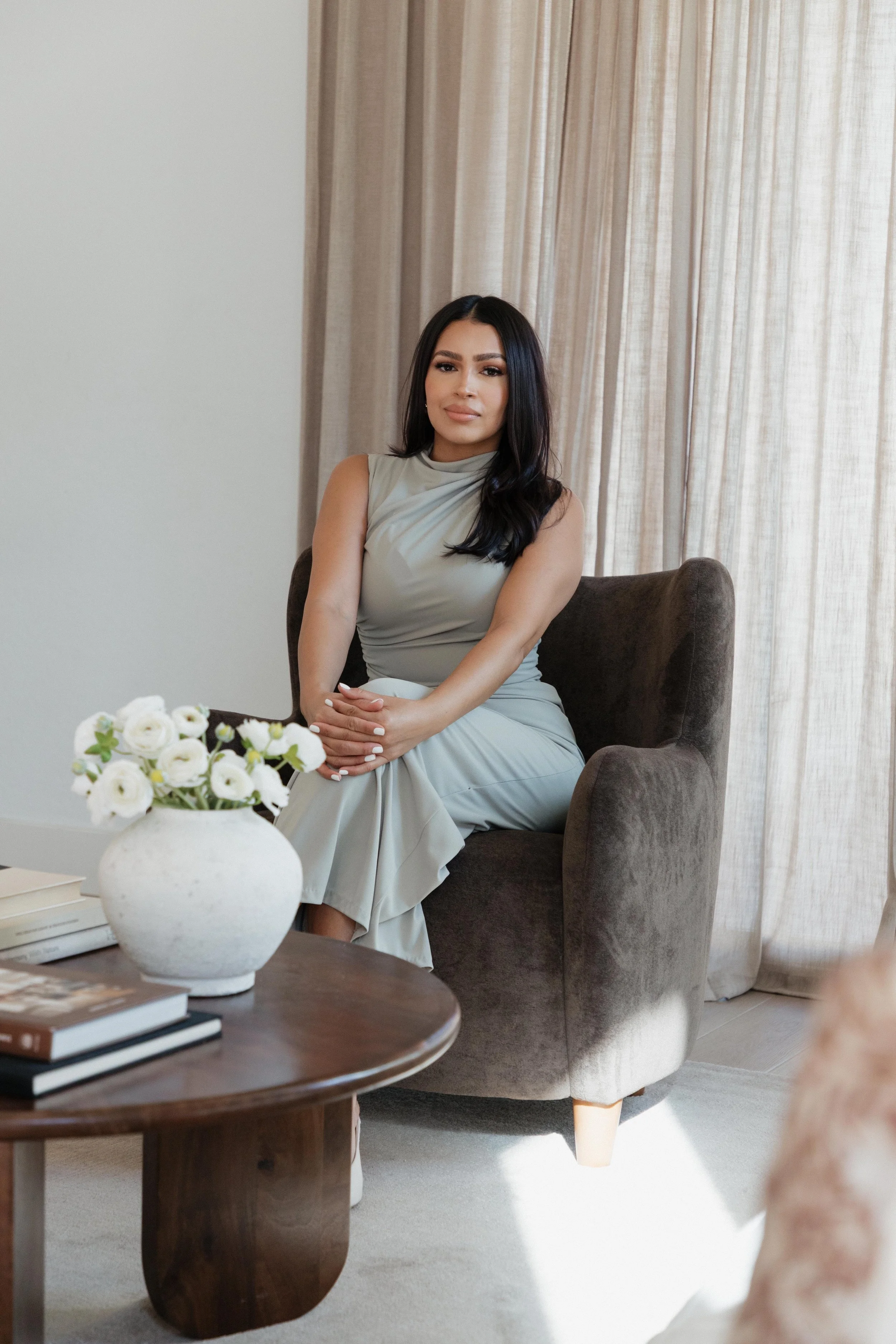 Alina Hernandez seated on a dark plum velvet armchair in a room with beige curtains. There is a white ceramic vase with white flowers on a wooden table in front of her.