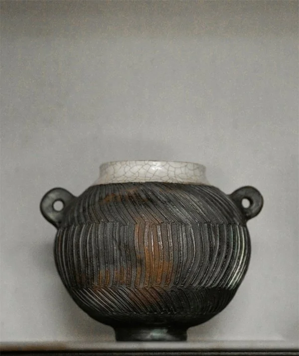 A round, dark-colored ceramic vase with two small handles and a white, crackled lid on top, displayed against a plain background.