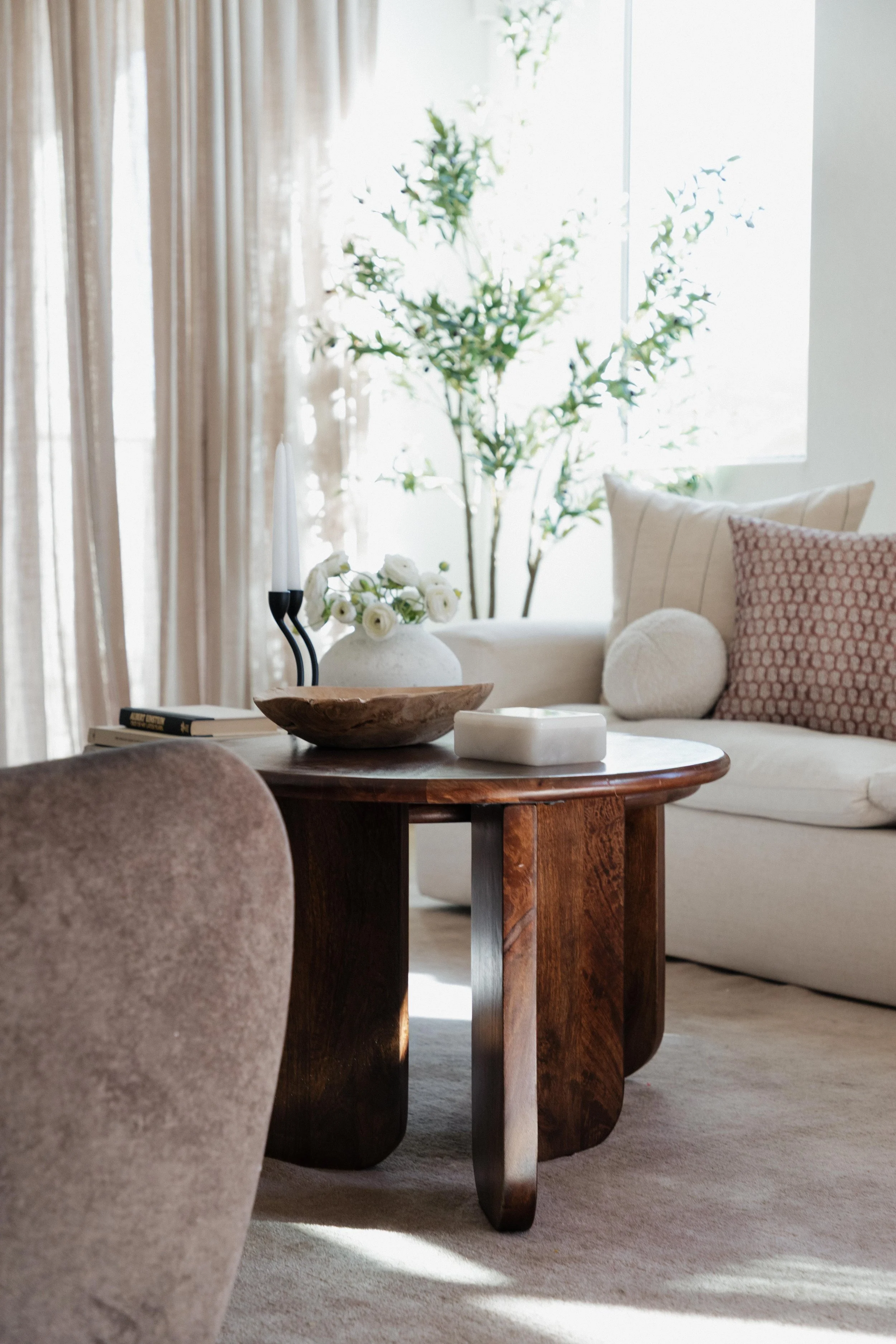 Living room interior with a wooden coffee table, beige sofa with pillows, a vase of white flowers, and a large window with curtains and a green plant in the background.