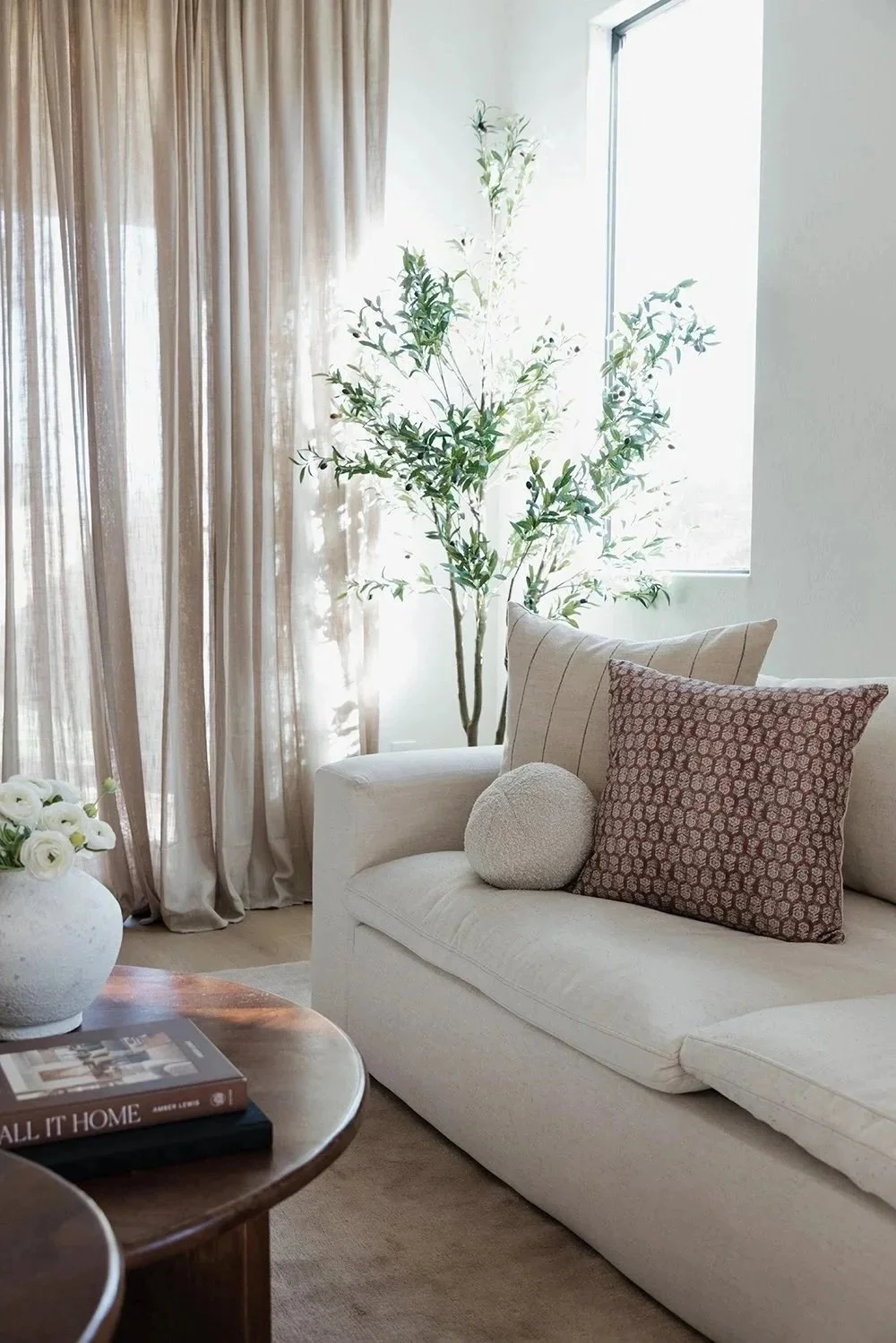 Living room with beige curtains, a tall indoor tree near a window, a cream sofa with decorative pillows, a wooden coffee table with books and a white vase with flowers.