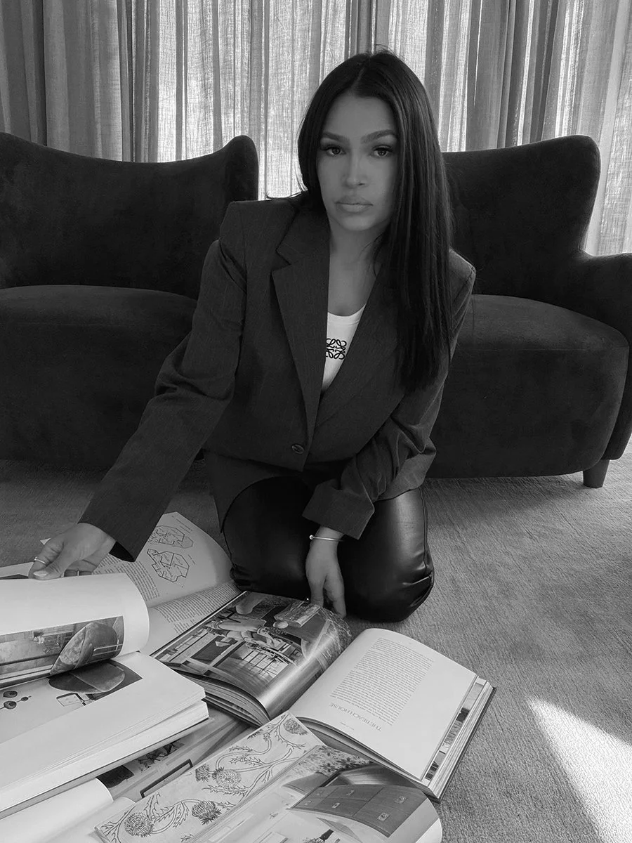 Black and white photo of a woman in a blazer kneeling on the floor, looking at open design and architecture books in a room with curtains and a sofa.