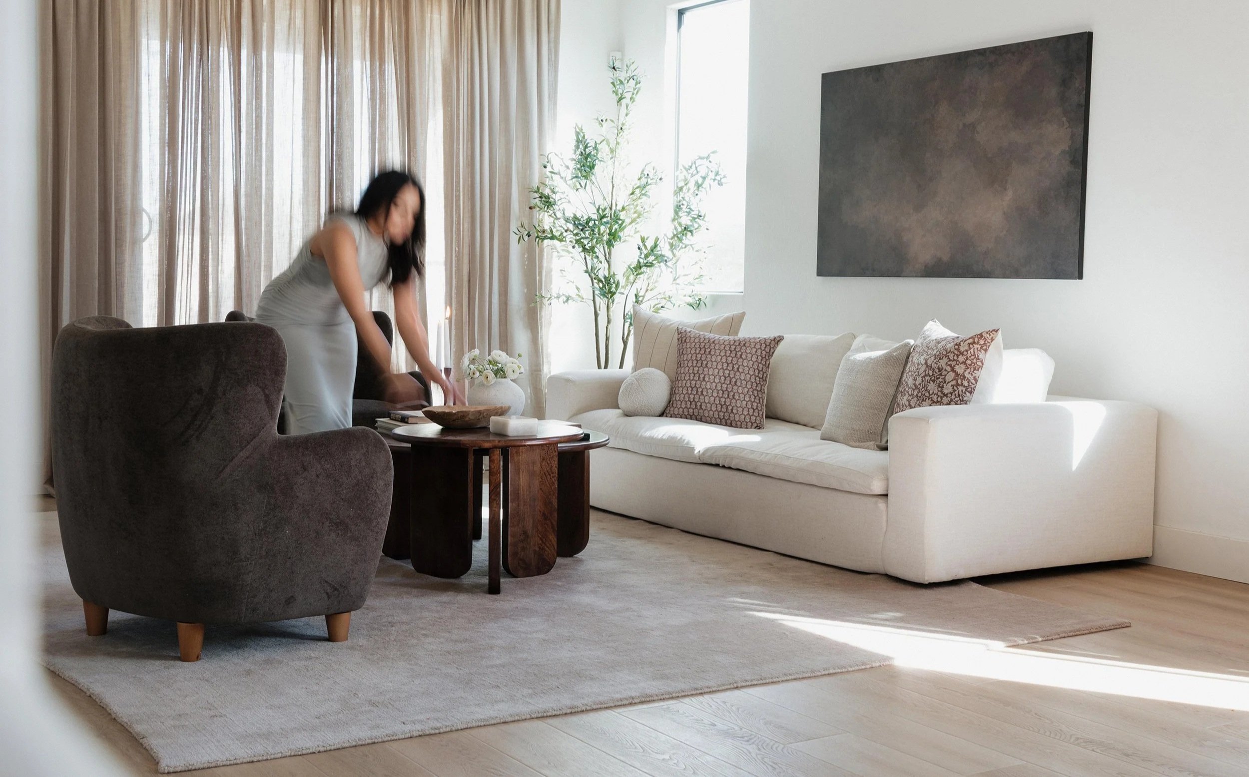 Modern living room with white sofa, dark armchair, round wooden coffee table, and beige area rug. Behind the sofa are beige curtains, a large abstract painting, and a window with sunlight. A woman is arranging items on the coffee table.