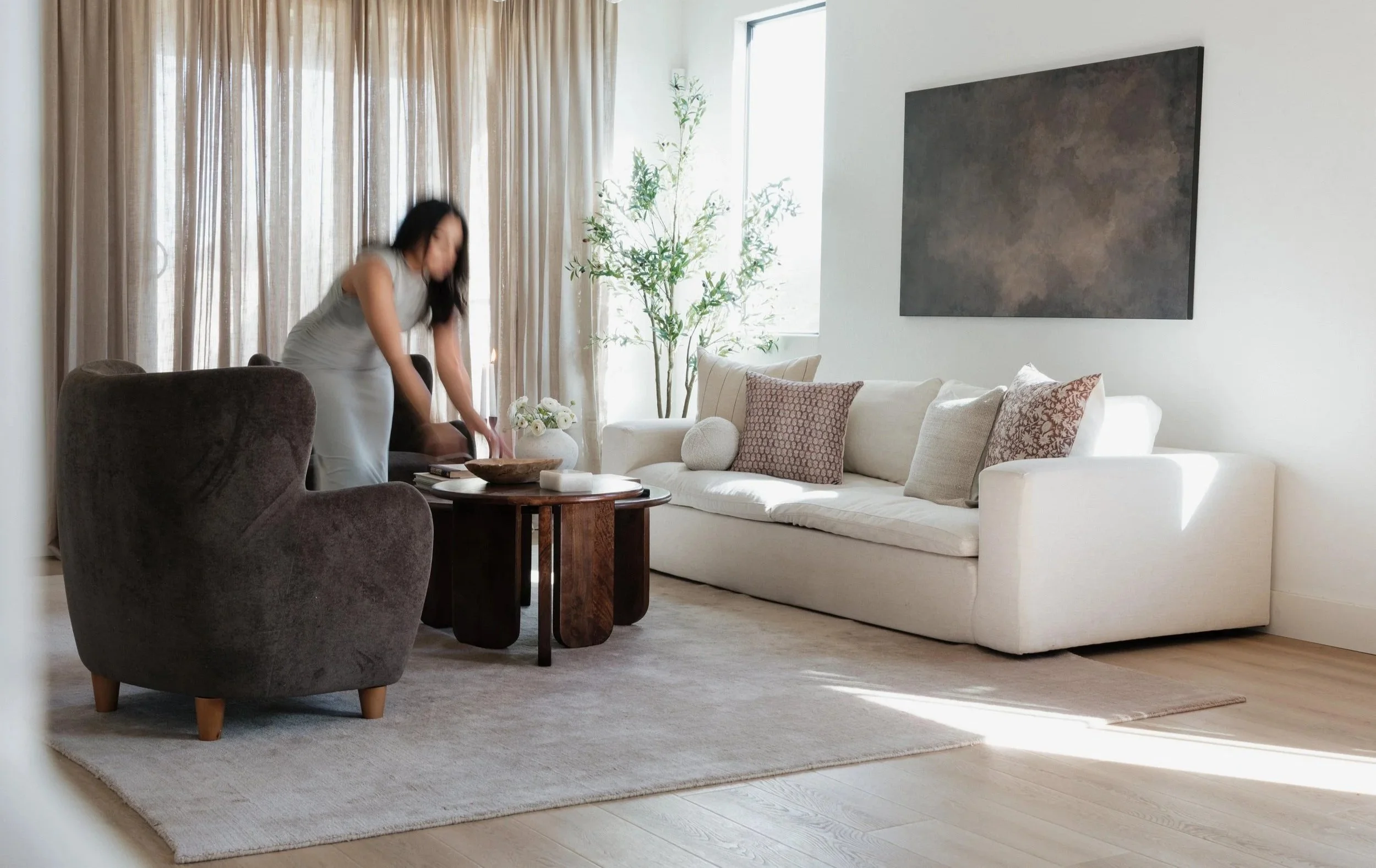 Interior designer works in an elegant modern living room with a white sofa, brown armchair, and large window with beige curtains. There is a large abstract painting on the wall.