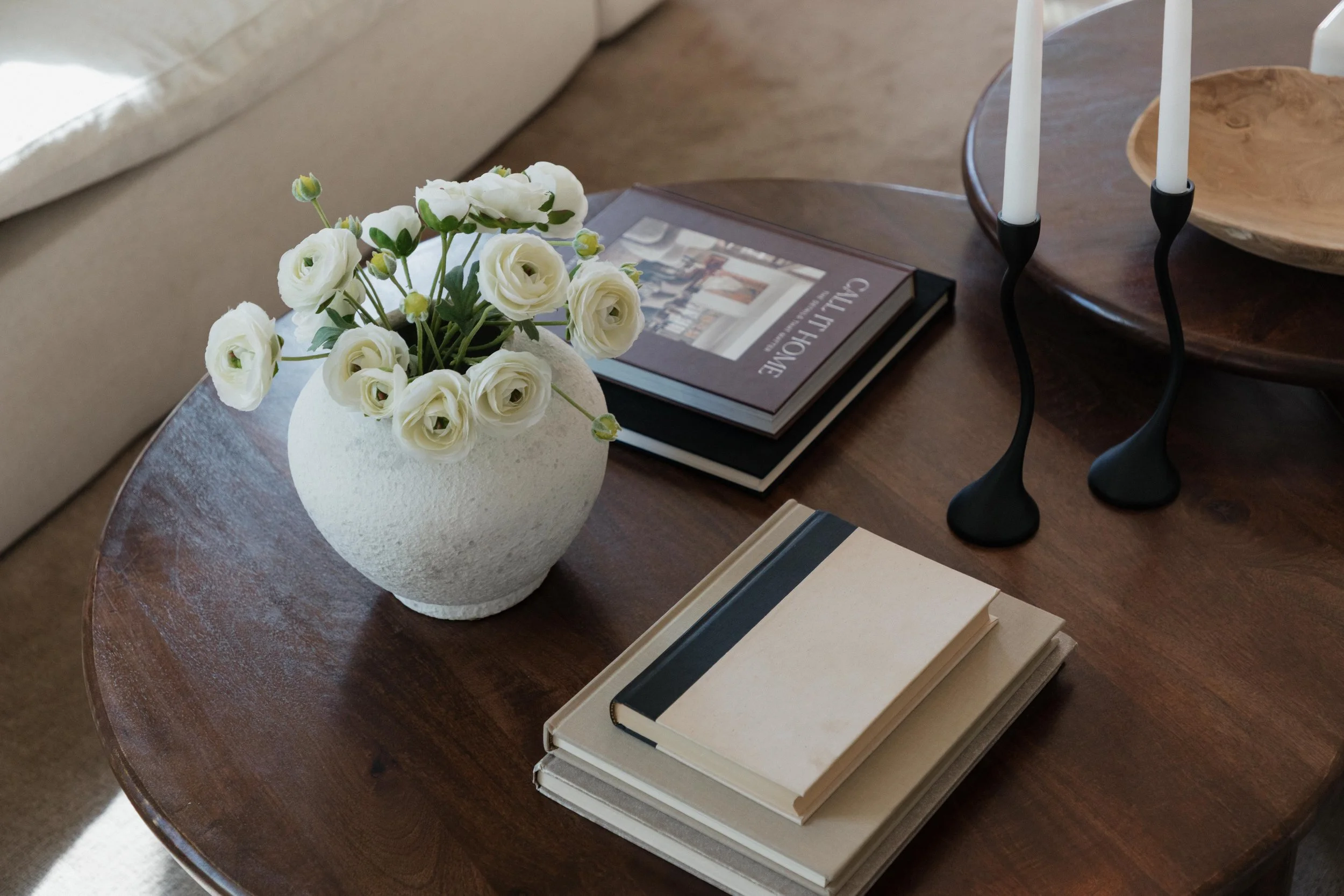 A round wooden table with a white ceramic vase containing white ranunculus flowers, a book titled 'Gallery Home,' a black and beige notebook, and a stack of books. There are also decorative black candle holders with white candles and a wooden bowl on the table.
