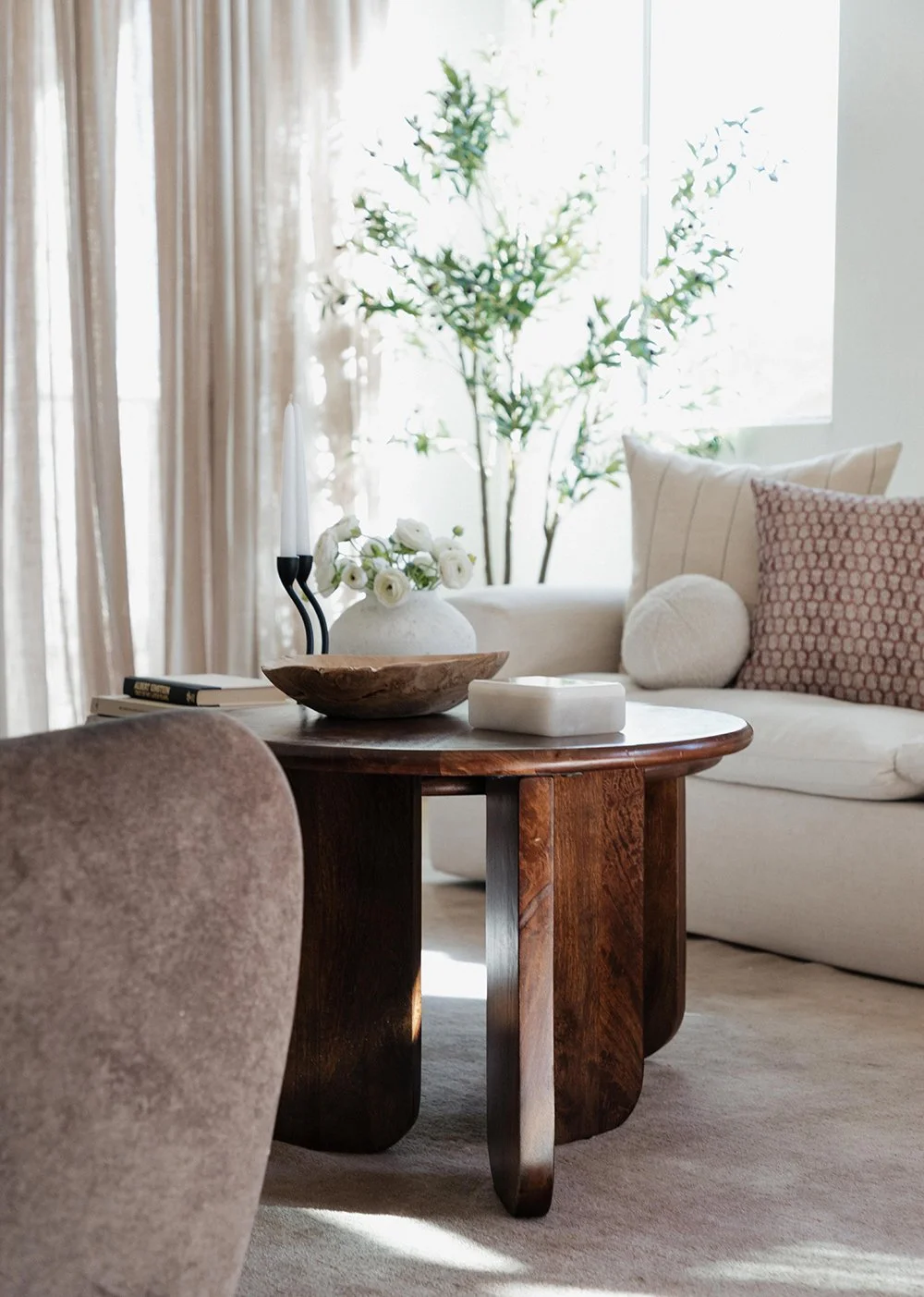 Living room with beige curtains, a white sofa with patterned pillows, a wooden coffee table with decorative items, and a large green plant near a window, illuminated by daylight.