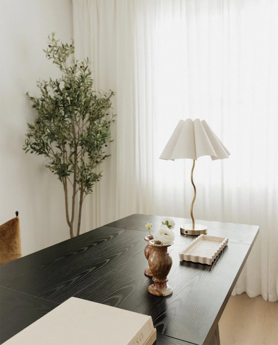 Black desk with a white flower vase, a beige tray, and a modern table lamp with a curved gold stand and a pleated white lampshade, in a bright room with cream curtains.