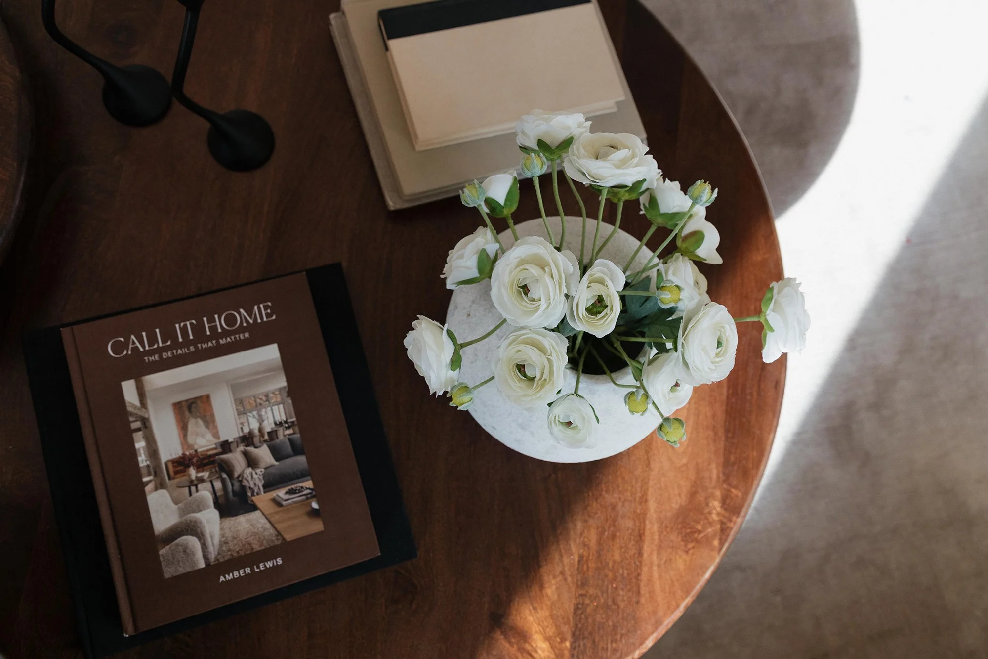 A wooden side table with a stack of interior design books, a black table lamp, and a white vase with white roses and buds.