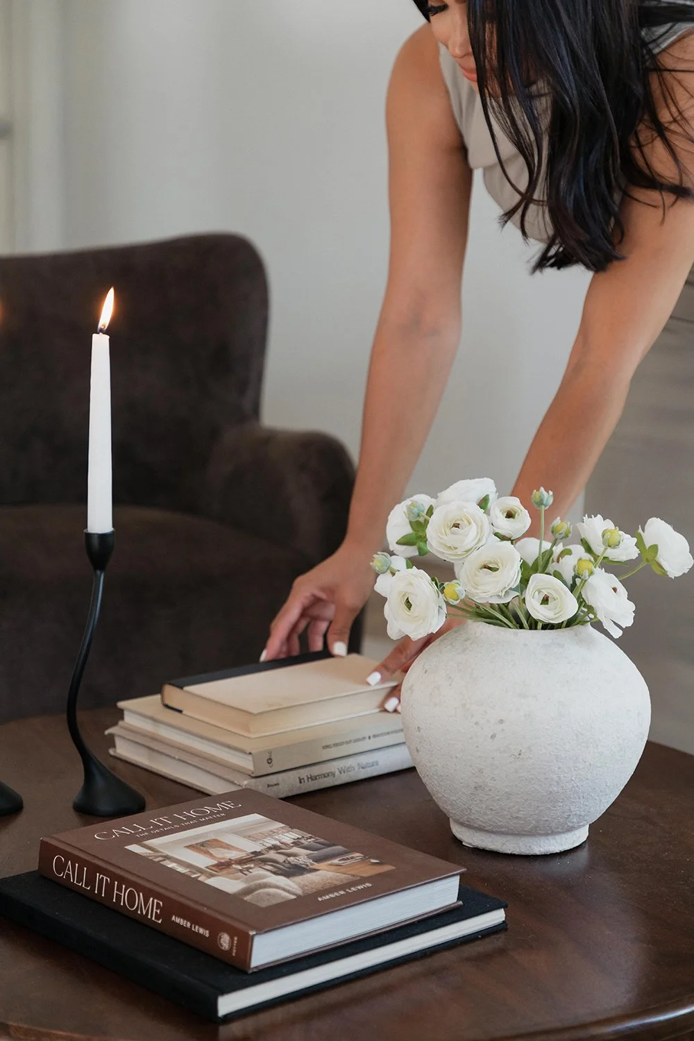 Interior Designer arranging books on a dark wooden coffee table. A white vase with white flowers, and a lit candle rest on the table in a cozy room.
