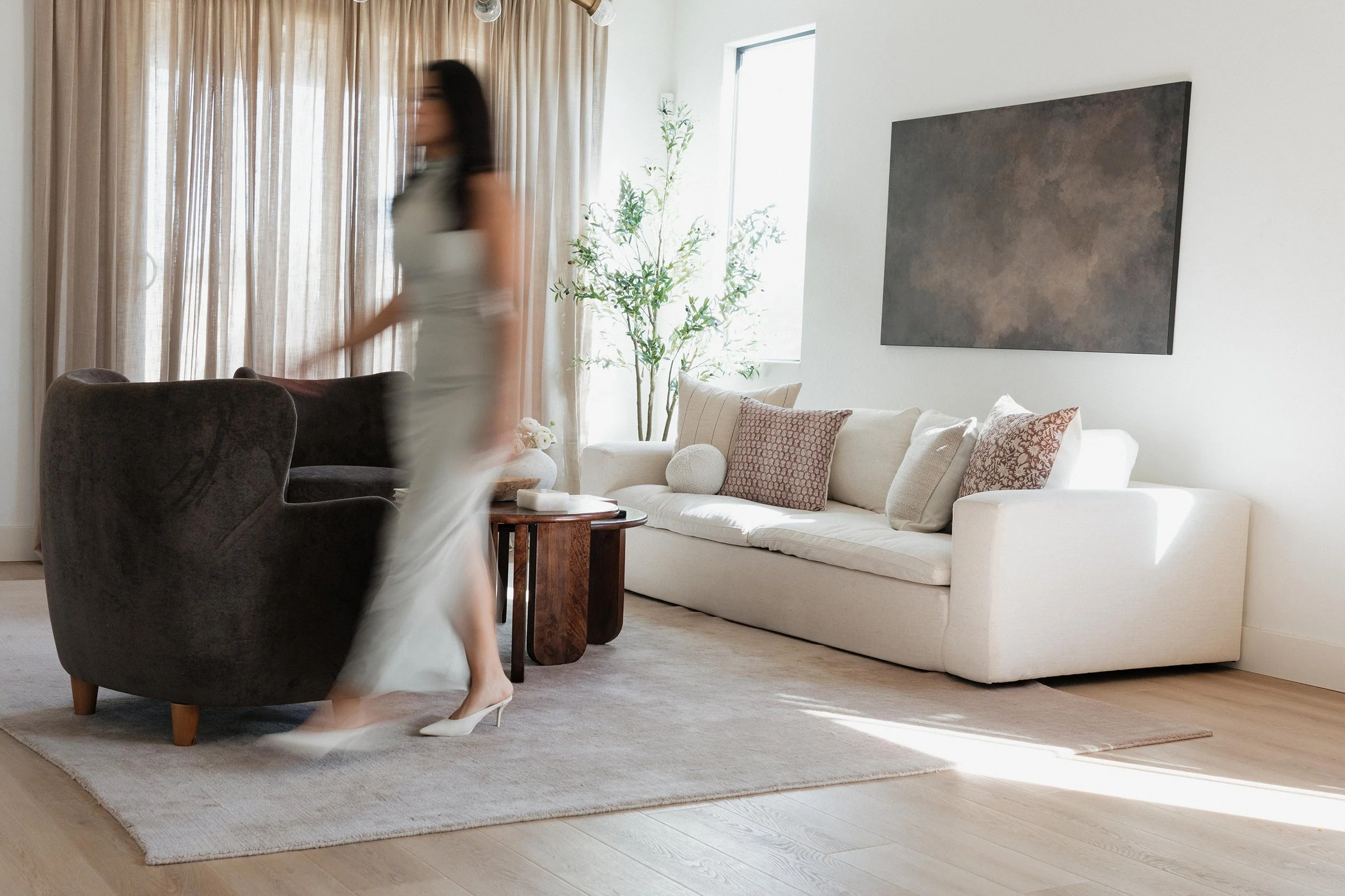 A woman with blurred motion walking through a bright living room with white sofa, patterned pillows, potted plant, and large window with beige curtains.