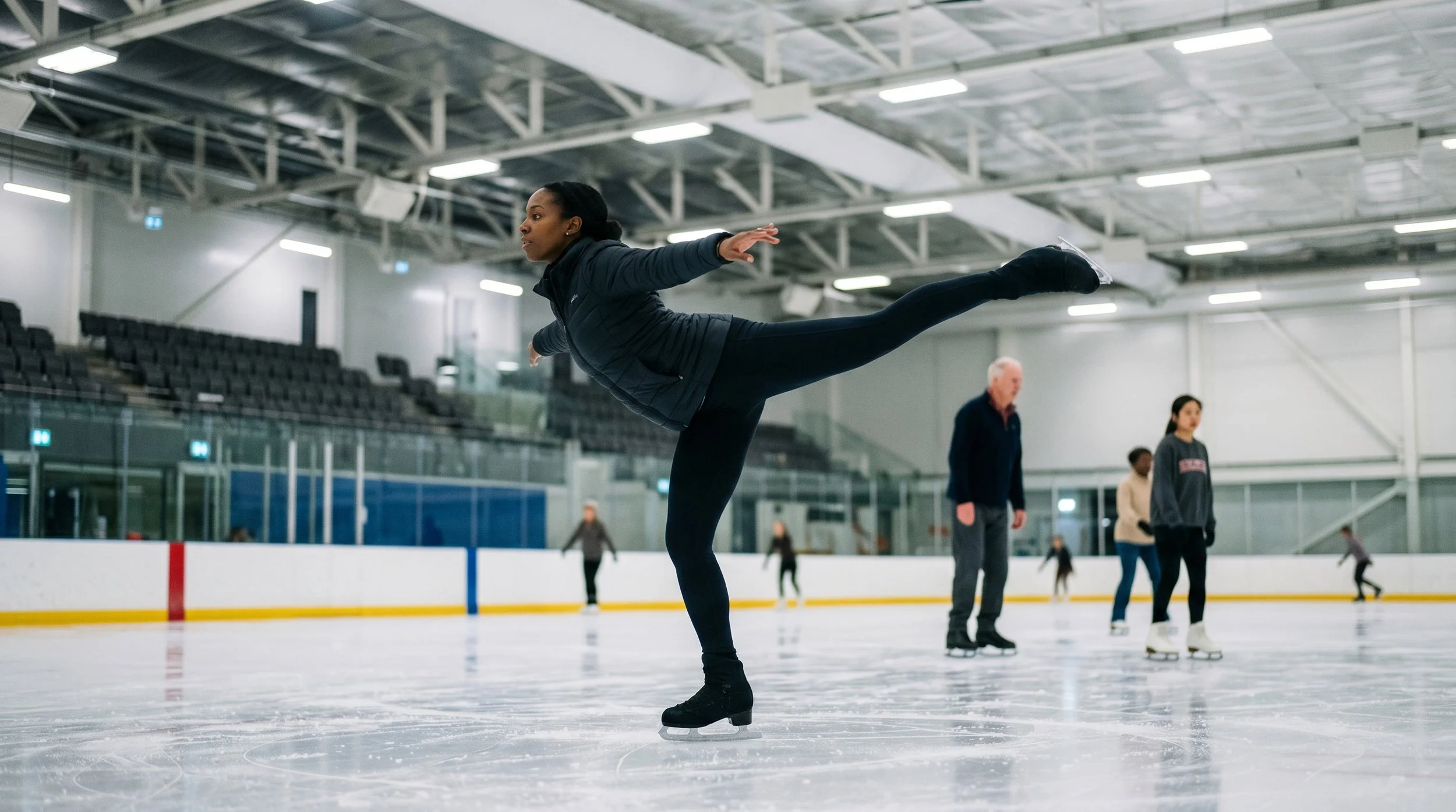 A woman ice skating on an indoor rink performing an advanced glide on 1 foot as other skaters move about in the background on an indoor ice rink