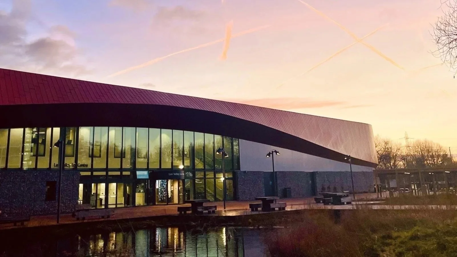 Lee Valley Ice Centre, A modern building with large glass windows and a curved roof, set against a sunset sky with pink and purple hues, trees without leaves, and a calm body of water reflecting the building and sky.