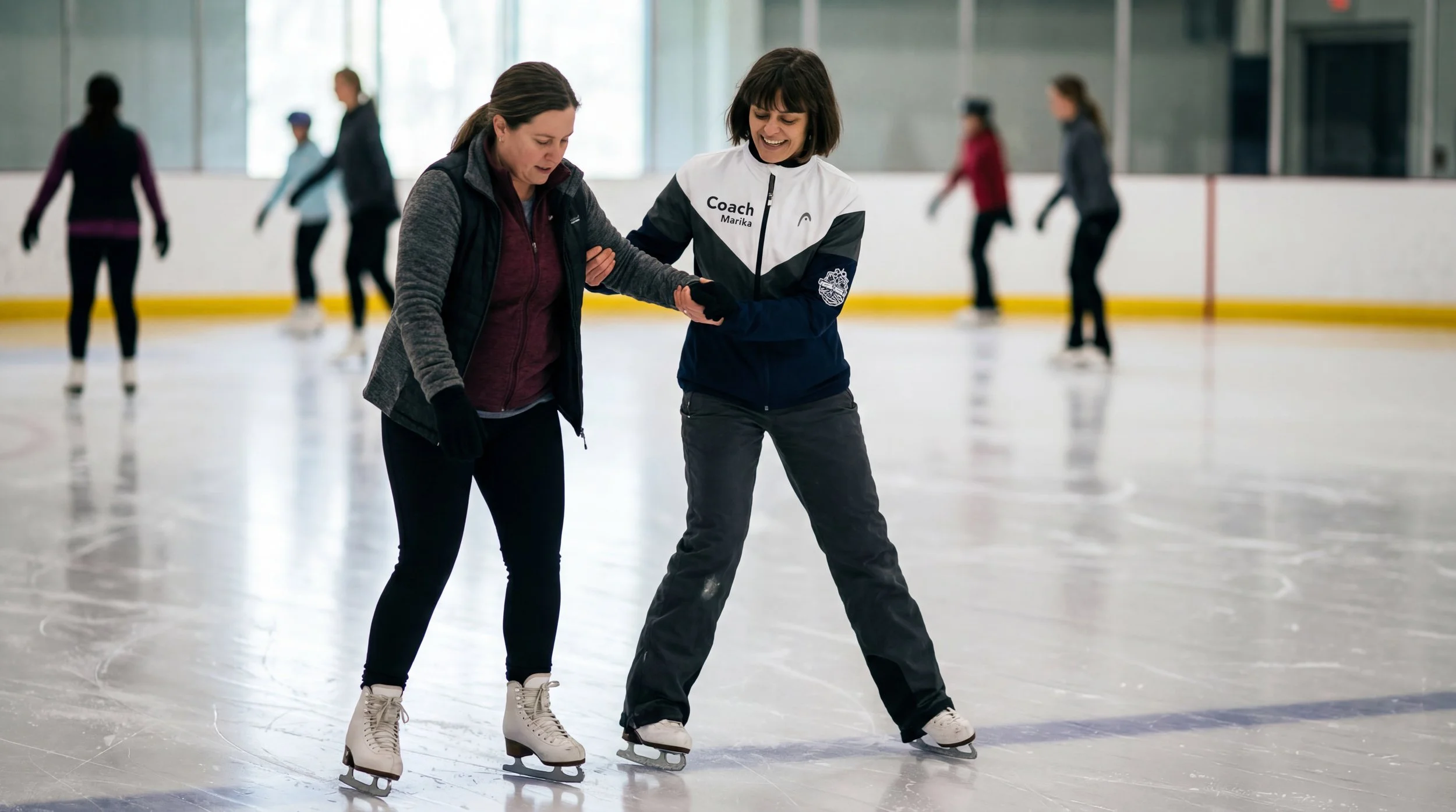A coach and a woman beginner ice skater are ice skating at an indoor rink, with others skating in the background.
