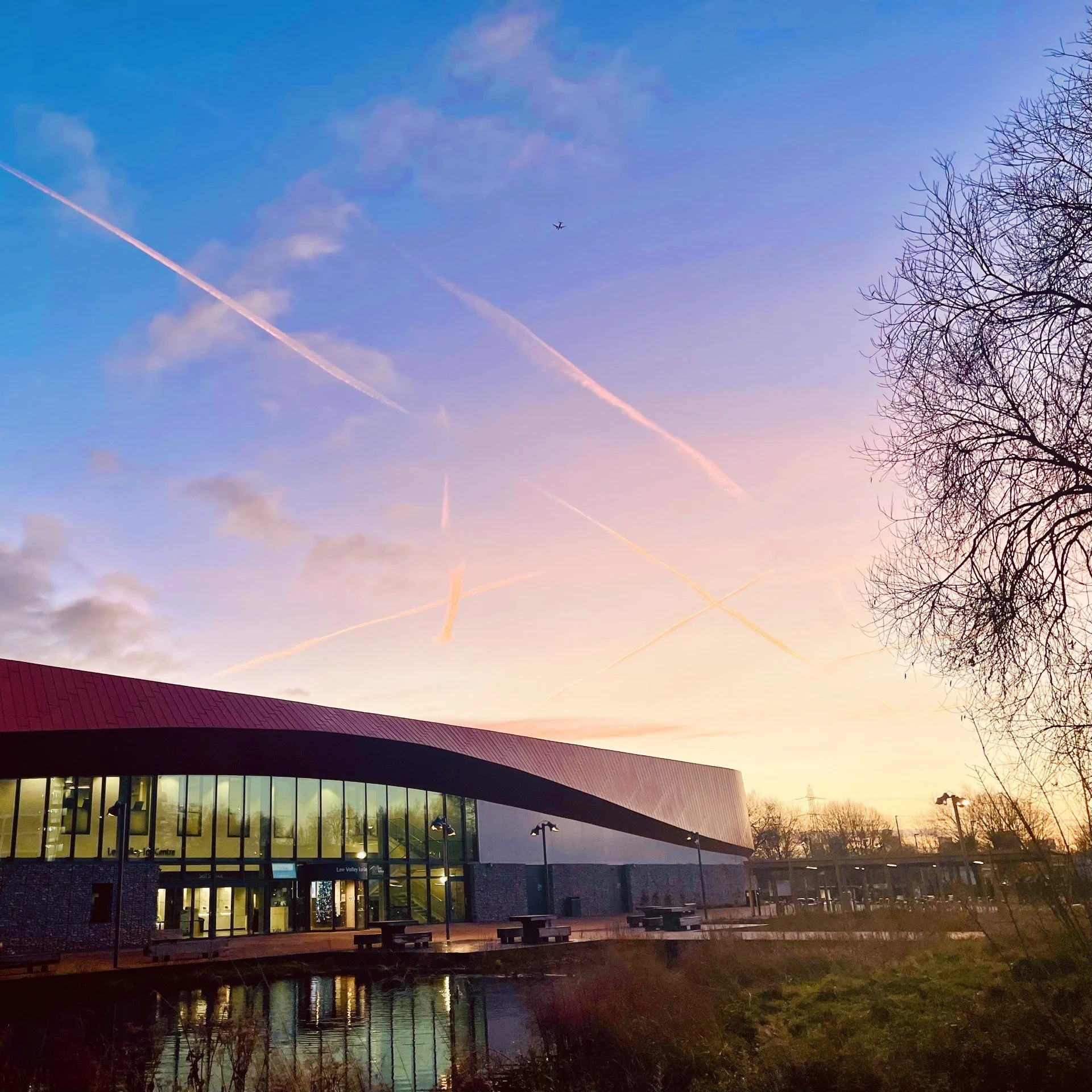Lee Valley Ice Centre, A modern building with large glass windows and a curved roof, set against a sunset sky with pink and purple hues, trees without leaves, and a calm body of water reflecting the building and sky.