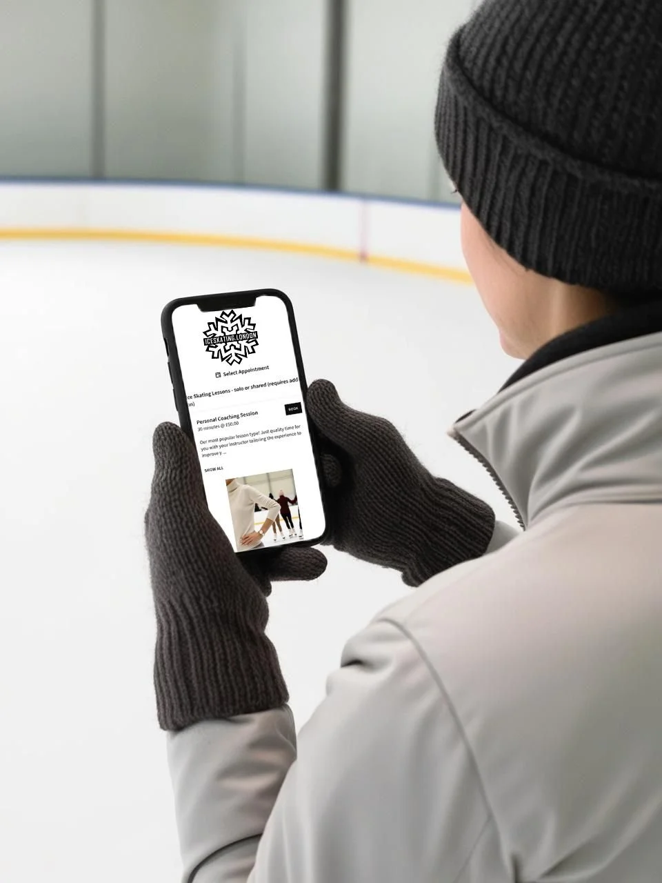 Person in winter clothing, including a gray jacket, black gloves, and a black knit hat, looking at a phone screen inside an ice skating rink.