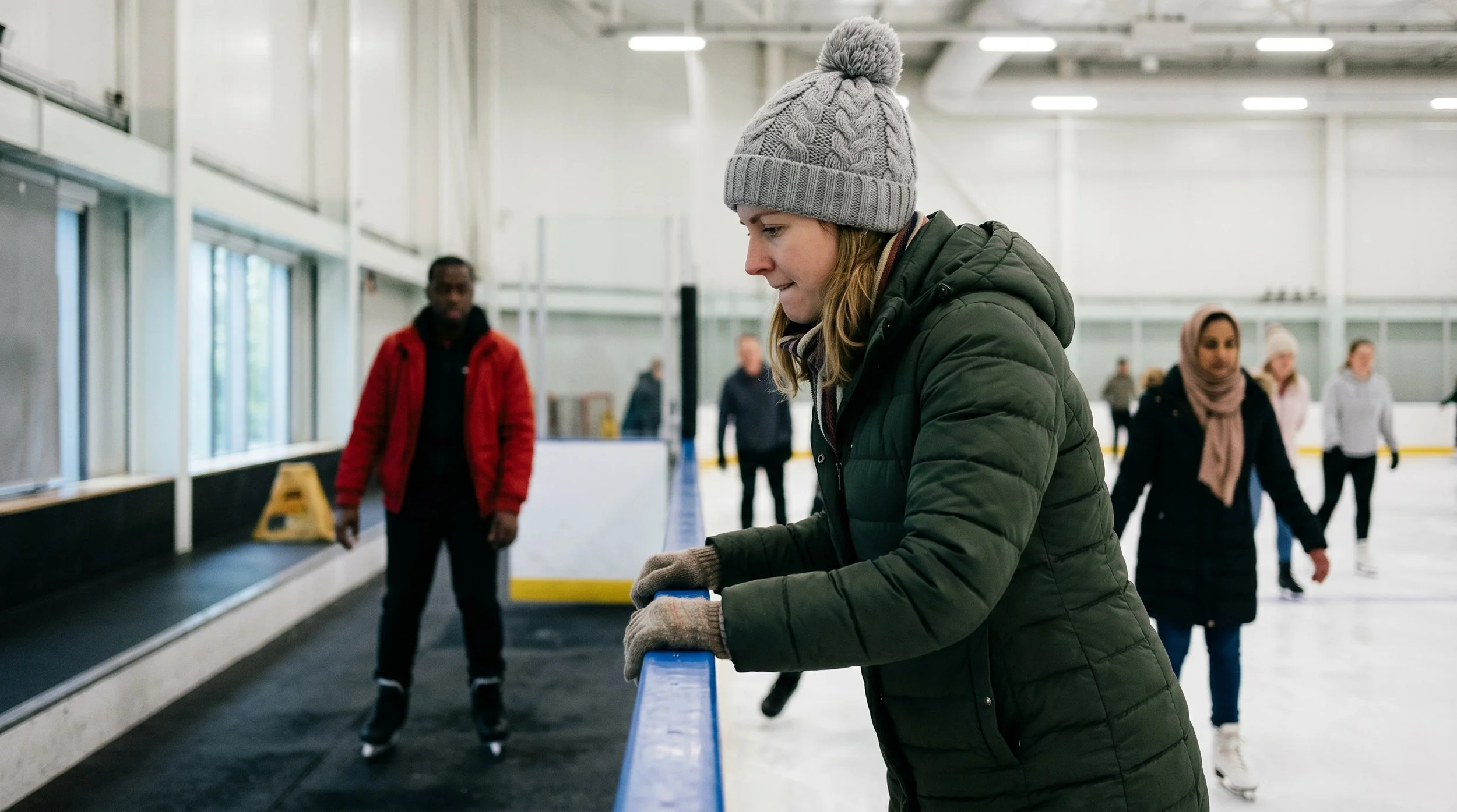 Woman wearing a grey cable knit bobble hat and a green down coat holds the side on an indoor ice rink barrier with an expression of mild anxiety