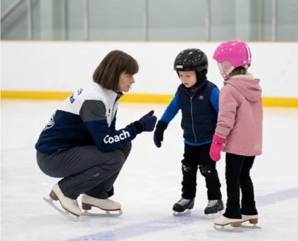 Ice skating coach instructing two children on ice rink.