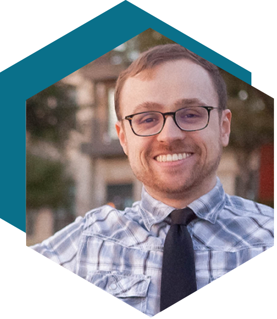 Smiling person with glasses wearing a plaid shirt and black tie, outdoors with a softly blurred background, framed in a hexagonal shape