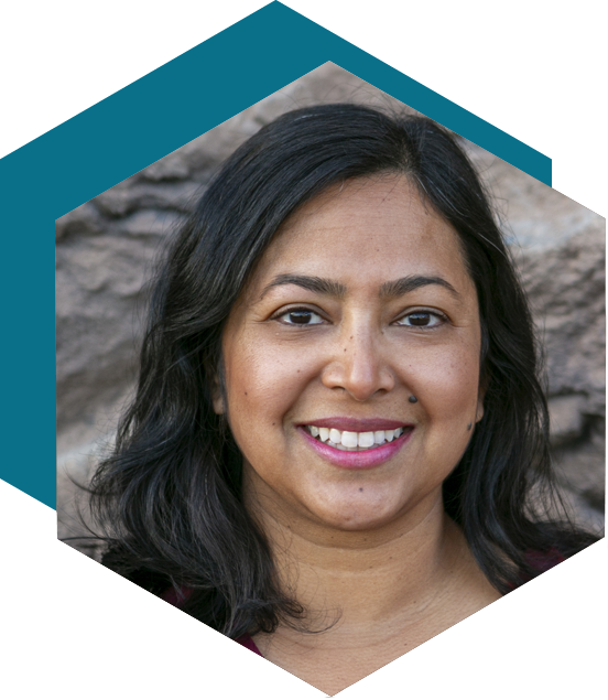 Smiling woman with shoulder-length dark hair in a maroon top, posed in front of a stone background, framed by a hexagon.
