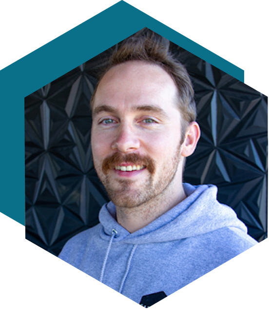 Smiling man with a mustache in gray sweatshirt with black shaped background within a hexagon