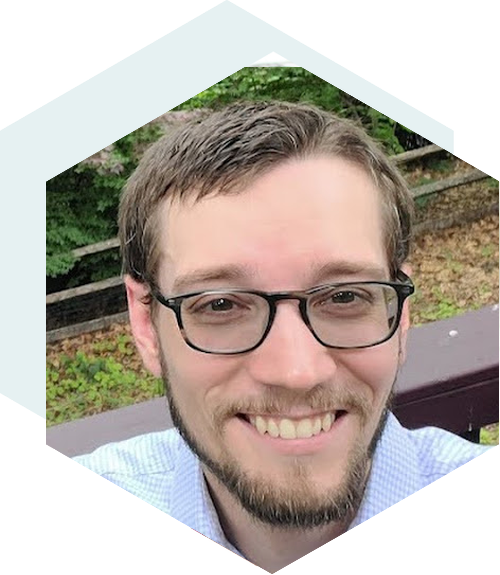 A smiling man with glasses and a beard, wearing a light blue shirt, outdoors with greenery in the background. The image is cropped into a hexagonal shape.