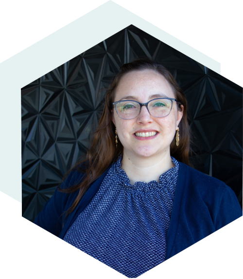 Smiling woman with long brown hair wearing glasses, a blue patterned top, and navy cardigan, standing in front of a black geometric wall, framed by a light hexagon shape