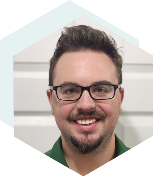 Man with curly hair and a beard, smiling in front of a brick wall, wearing a suit and tie, framed by a blue hexagon shape