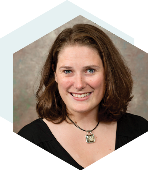 A smiling woman with shoulder-length brown hair, wearing a black top and a pendant necklace. The background is a neutral studio backdrop, and the image is cropped into a hexagonal shape.