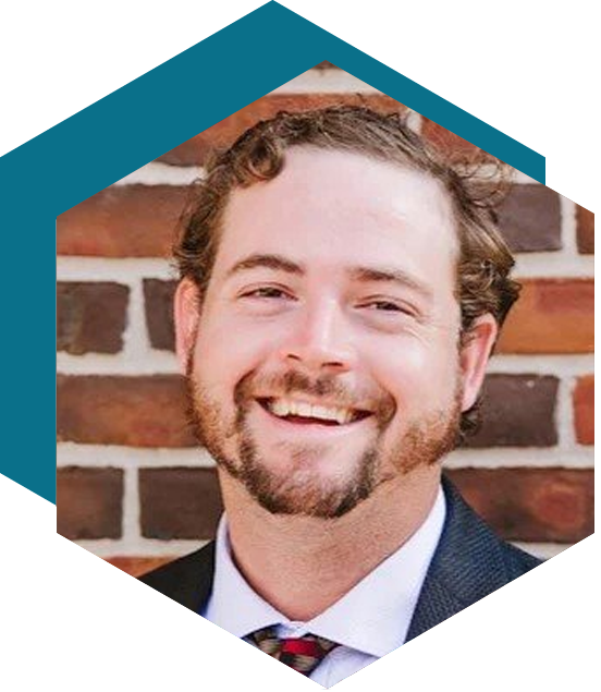 Smiling man in a suit with curly hair and beard, standing in front of a brick wall, framed by a blue hexagon graphic