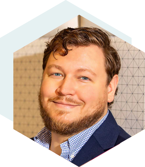 Smiling man with short curly brown hair and a beard, wearing a navy blazer over a checkered shirt, set against a patterned background with a hexagonal design overlay.
