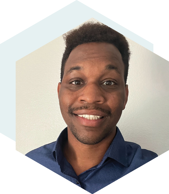 Smiling man with short curly hair, wearing a blue collared shirt, posing in front of a plain white wall with a hexagonal design in the background.
