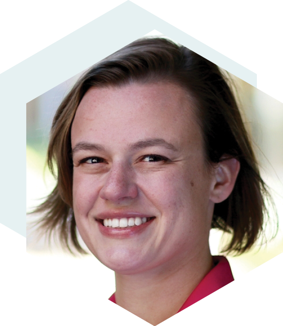 Smiling woman with short brown hair, wearing a pink collared shirt, set against a blurred outdoor background with a hexagonal design overlay.