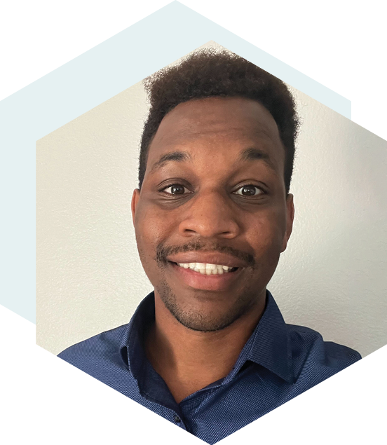 Smiling man with short curly hair, wearing a blue collared shirt, posing in front of a plain white wall with a hexagonal design in the background.