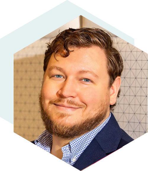 Smiling man with short curly brown hair and a beard, wearing a navy blazer over a checkered shirt, set against a patterned background with a hexagonal design overlay.