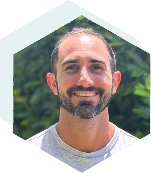 Smiling person with short dark hair and a trimmed beard wearing a light gray shirt, outdoors with greenery in the background, framed in a hexagonal shape.
