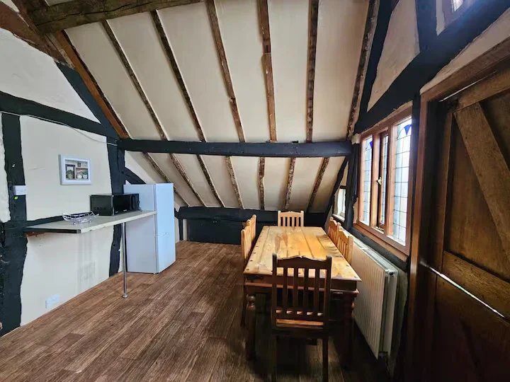 Attic room with sloped ceiling, wooden floor, fireplace, white refrigerator, microwave, and a dining table with four chairs near windows.