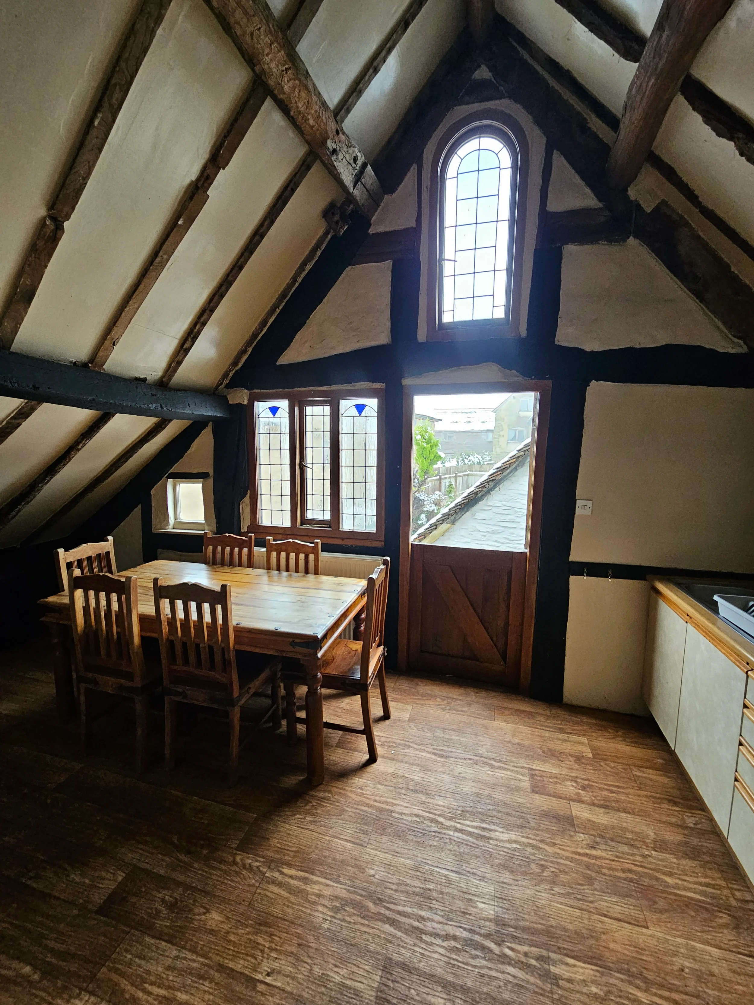 A rustic-style dining area featuring a wooden table surrounded by six matching chairs, with large windows that allow natural light to fill the space, a sloped ceiling with exposed dark wooden beams, and a view of neighboring houses and a tree outside.