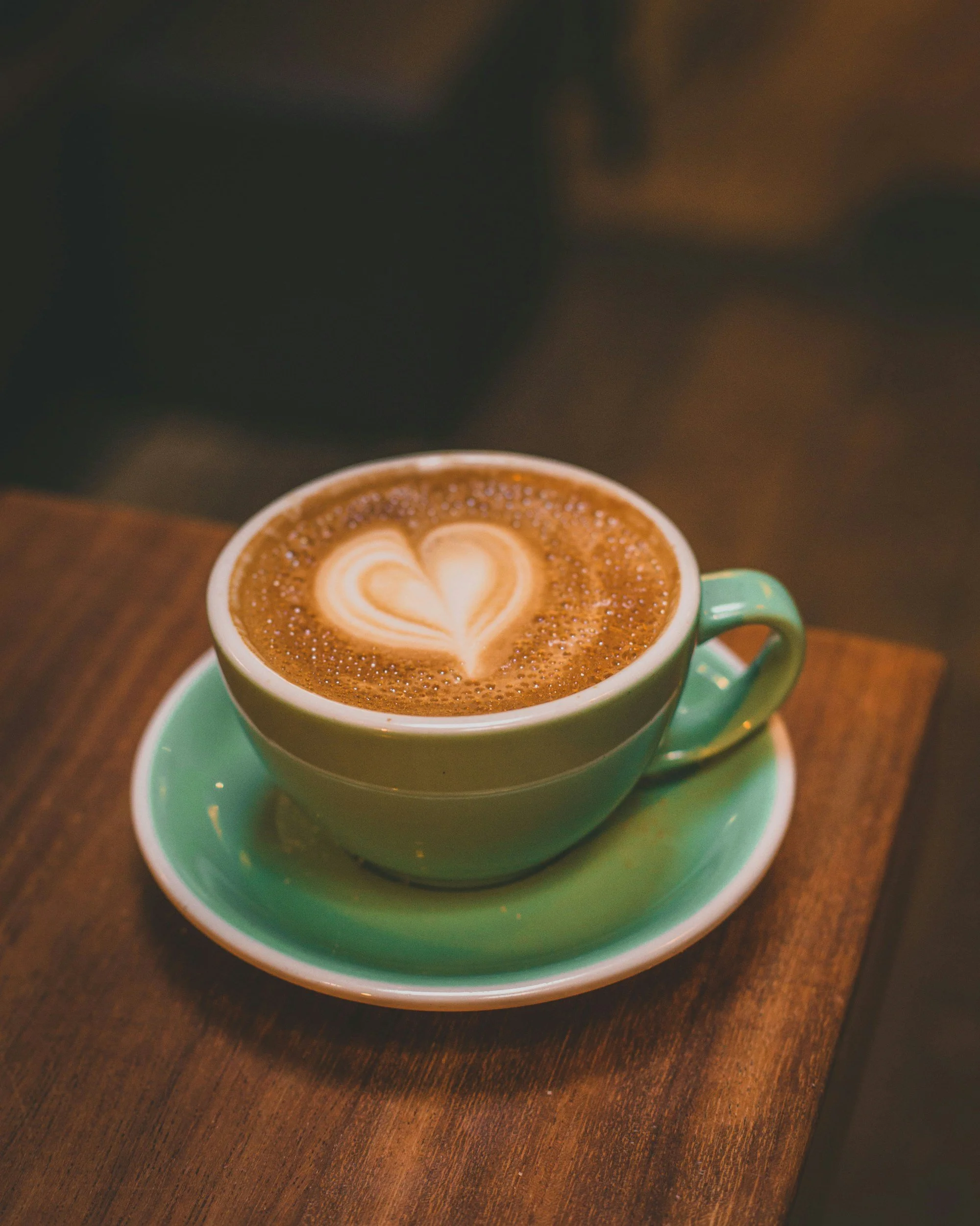 A green cup of hot chocolate with heart latte art, on a matching green saucer, placed on a wooden table.