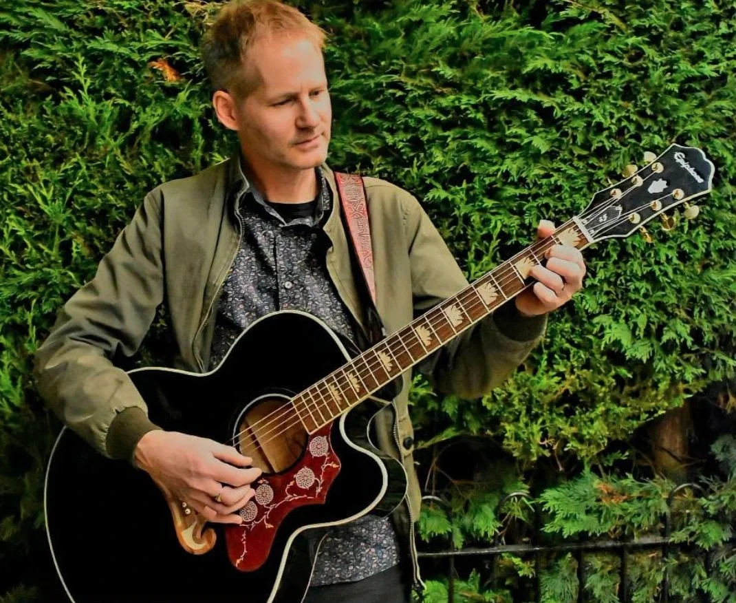 A man playing a black acoustic guitar outdoors with green foliage in the background.