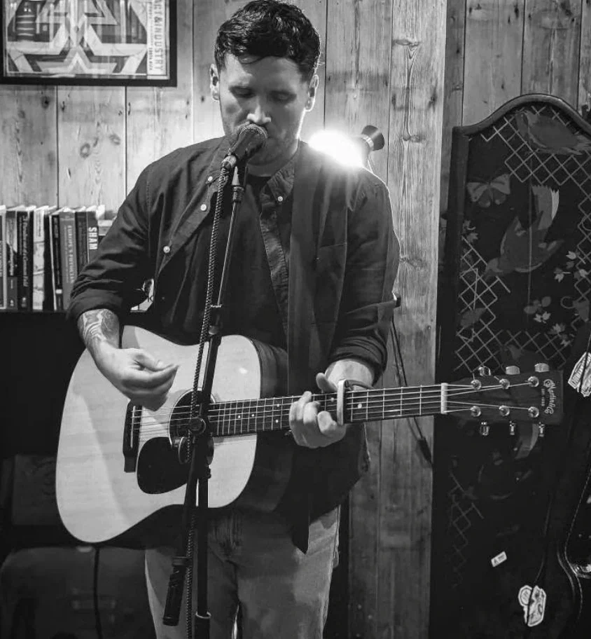 A man with dark hair playing an acoustic guitar and singing into a microphone in a cozy, wooden-paneled room, with books on a shelf and an acoustic guitar case in the background.