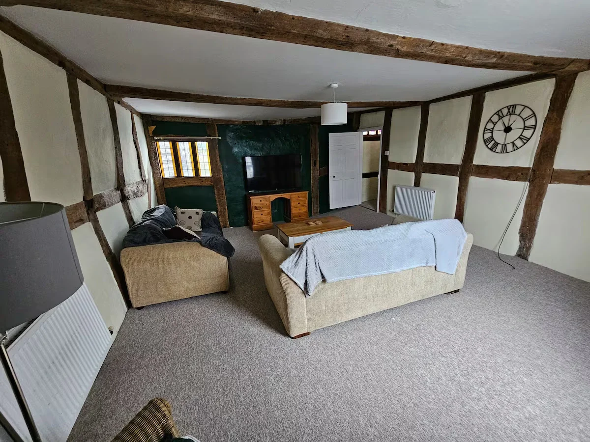 Living room with exposed wooden beams, a beige sofa with a white throw, a smaller beige armchair, a flat-screen TV on a wooden stand, a coffee table, a wall clock, and a radiator.