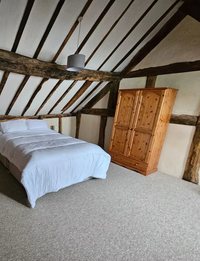 A bedroom with a sloped ceiling featuring exposed dark wooden beams, a bed with white bedding, a wooden wardrobe, a small bedside lamp, and a grey lampshade hanging from the ceiling.