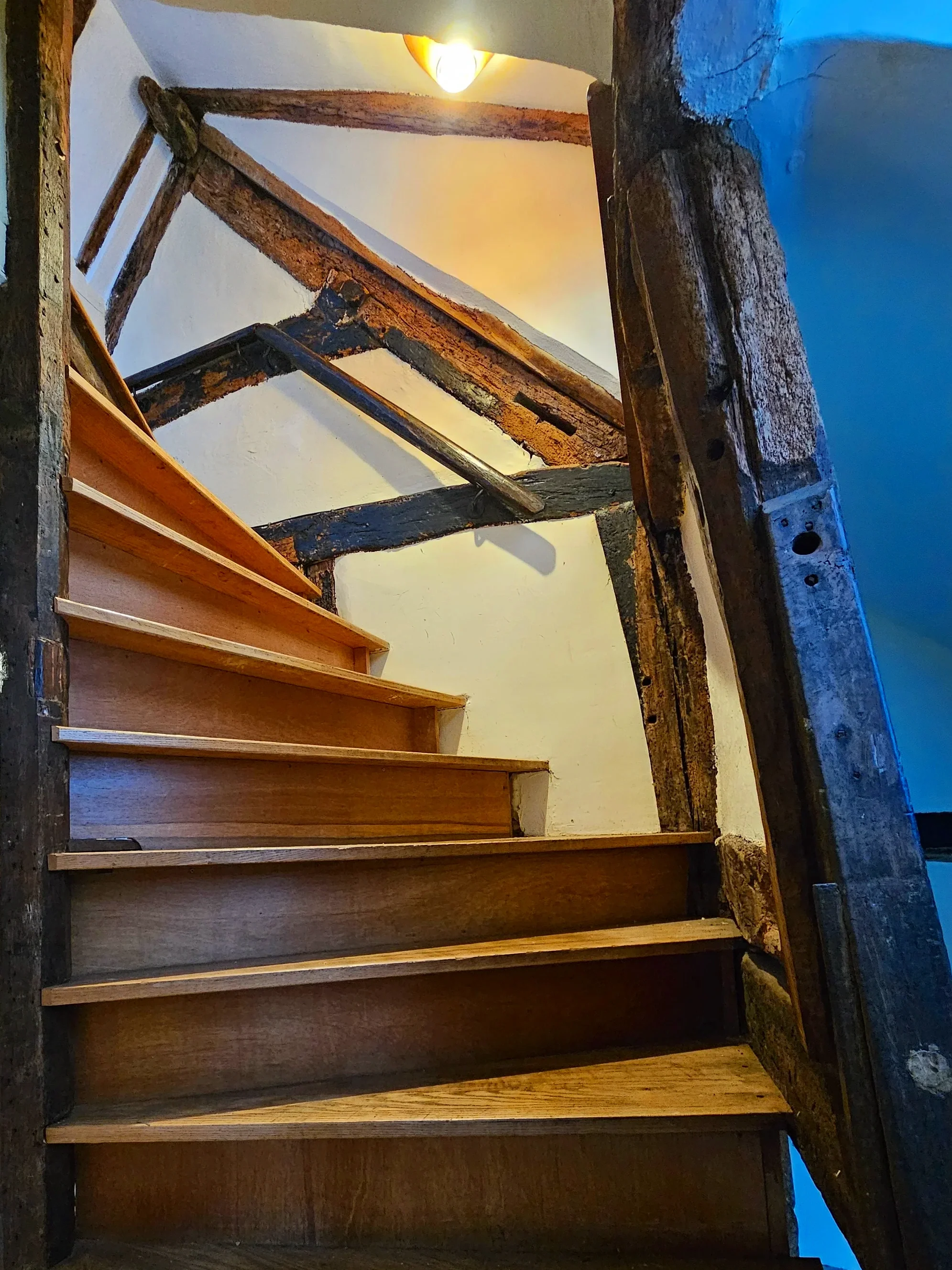 Interior view of wooden stairs with unfinished and weathered wooden railing, under a ceiling light fixture, with a wall painted in addition to exposed wooden beams.