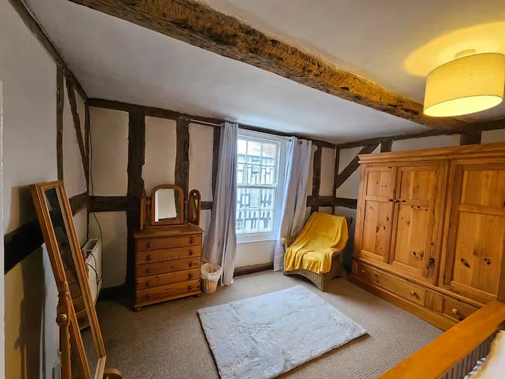 A cozy attic bedroom with sloped ceiling and exposed wooden beams, featuring a wooden dresser with mirror, a yellow armchair with a blanket, a large wooden wardrobe, a window with white curtains, and a small rug on carpeted floor.