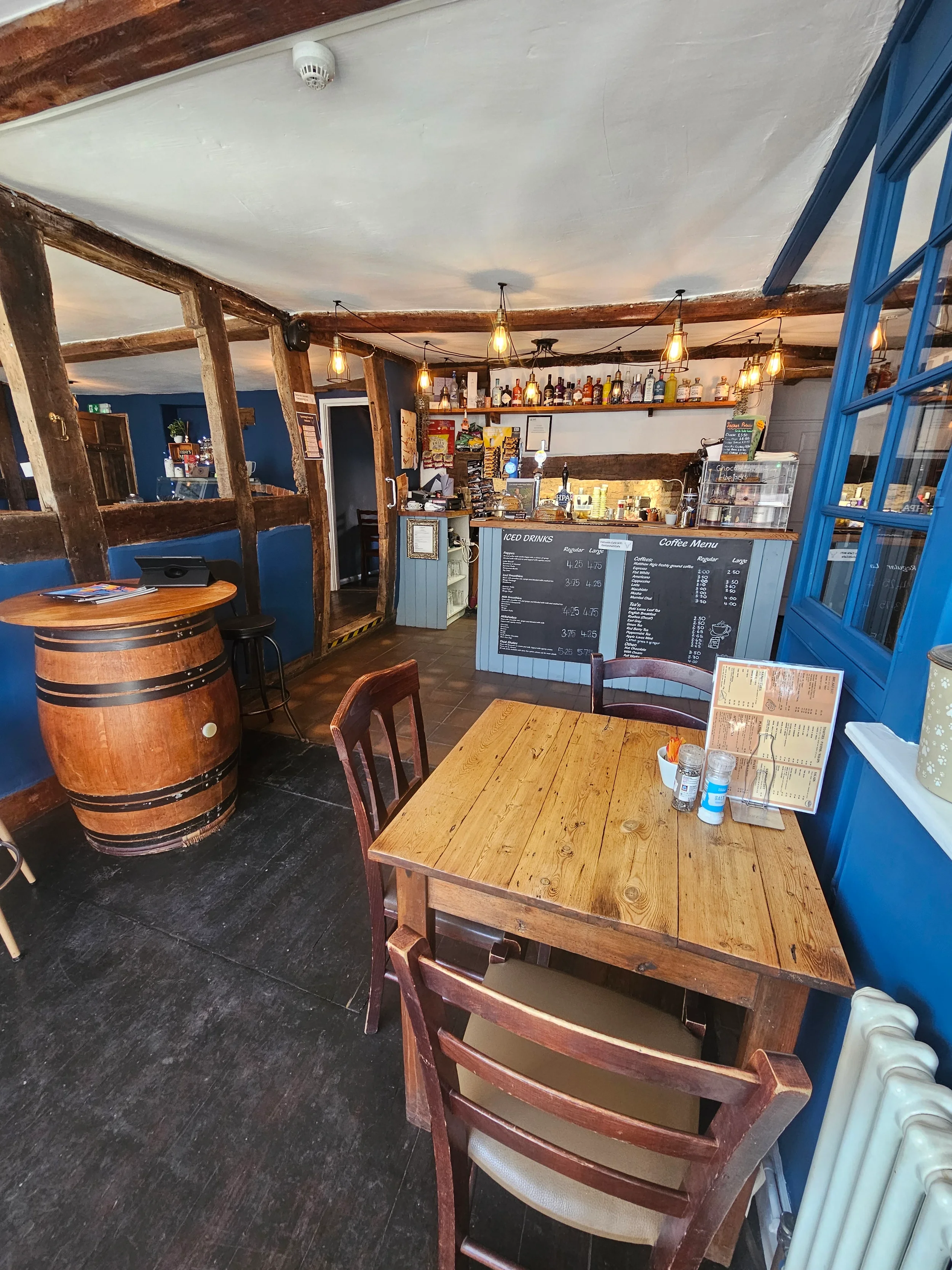 Interior of a cozy cafe with wooden beams, tables, and chairs, a bar with bottles, chalkboard menus, and warm lighting.