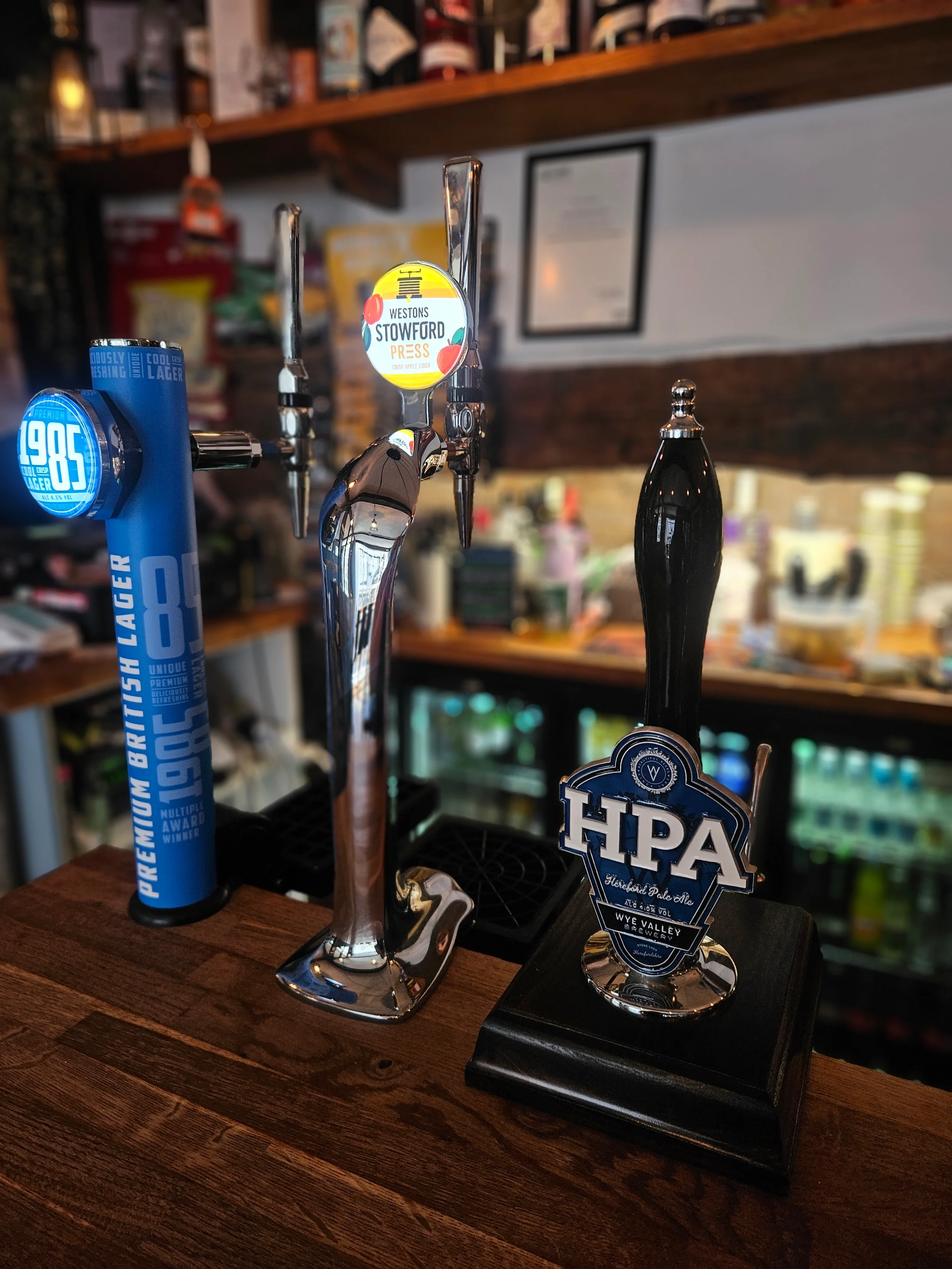 A collection of beer taps on a wooden bar counter, with various labels and a foam stand at a brewery or bar setting, with shelves in the background.