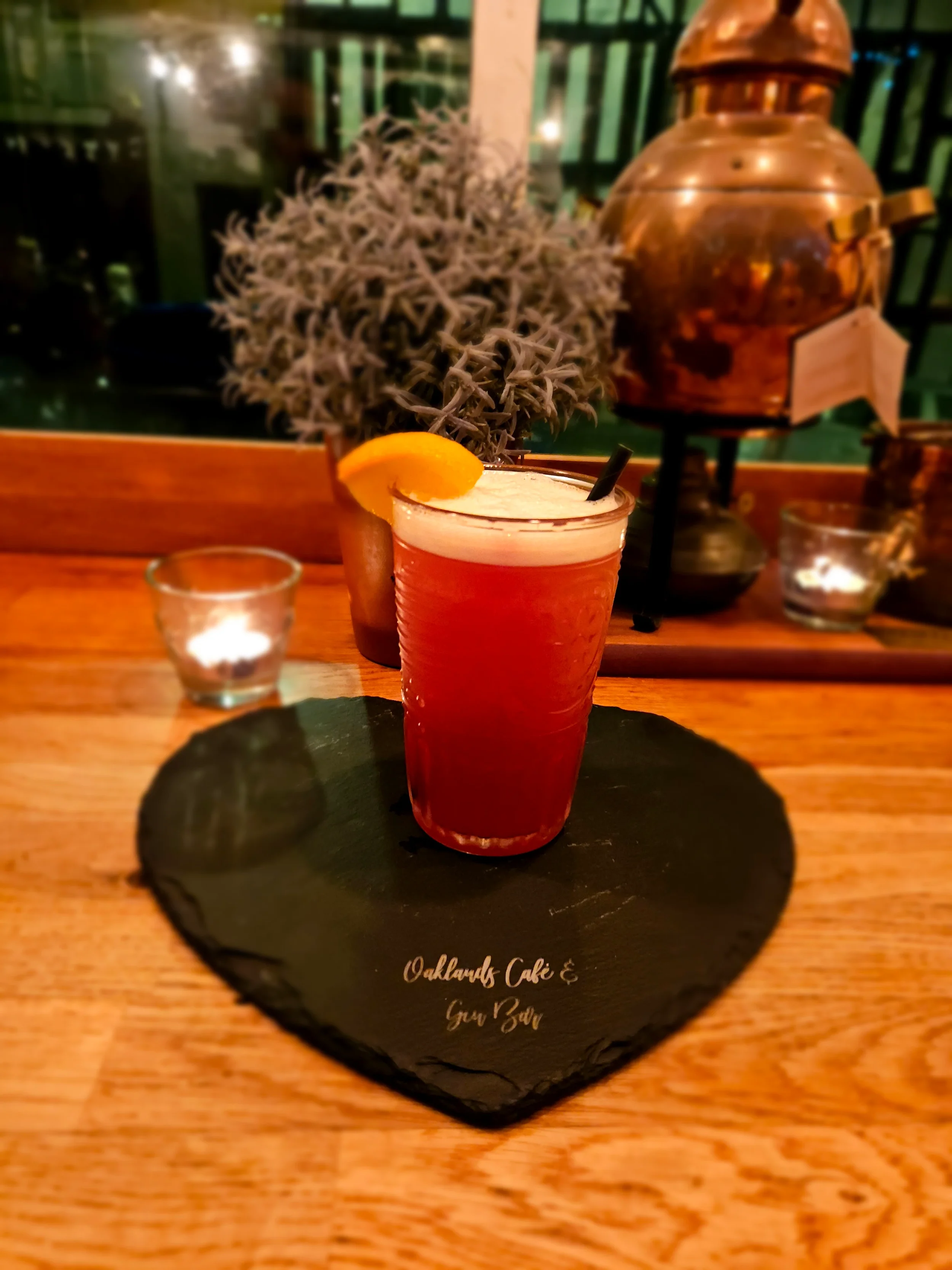 A glass of red cocktail garnished with a lemon slice on a black slate plate with the words "Oaklands Cafe & Bar" written on it. In the background, there is a decorative plant and a copper tea pot.